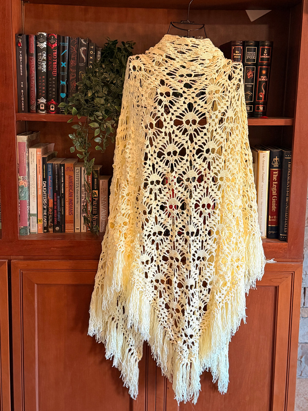 A cream-colored crocheted lace shawl with fringe trim hanging on a hanger in front of a wooden bookshelf filled with books.