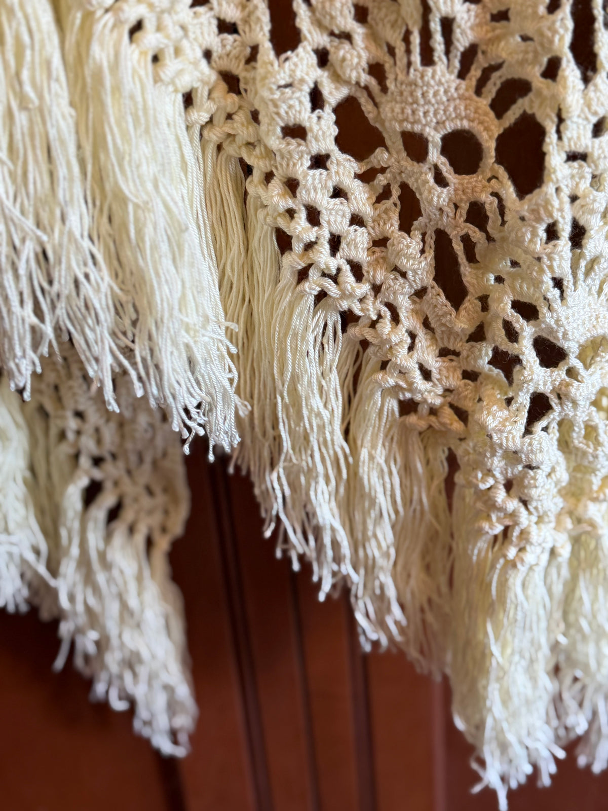 Close-up of a cream-colored crocheted or lace textile with long fringe tassels, draped over a dark brown wooden surface.